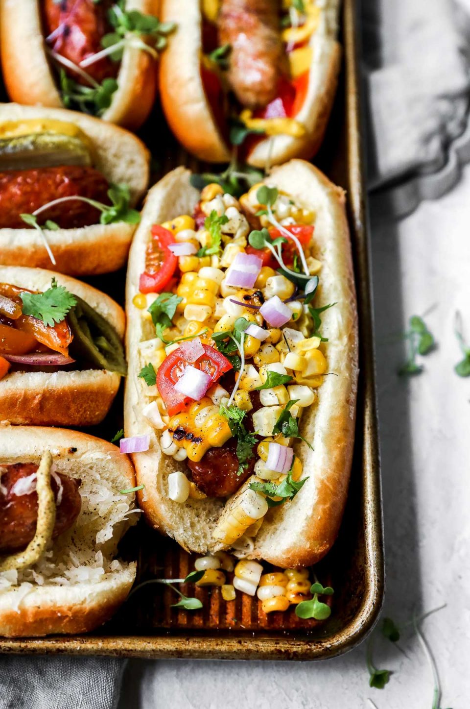Grilled sausage on a small baking sheet, served on a bun & topped with a fresh corn salad. The baking sheet is on a white surface, sitting atop a grey linen napkin. 