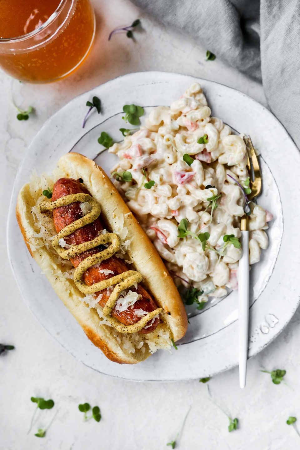 Grilled Sausage on a bun, topped with brown mustard. The sausage is plated with a simple macaroni salad on the side, on a white plate with a gold & white fork. 
