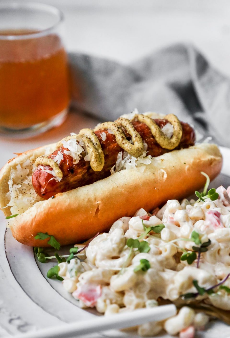 Side angle of Grilled Sausage on a bun, topped with brown mustard. The sausage is plated with a simple macaroni salad on the side, on a white plate with a gold & white fork. Off in the background, there is a glass of beer & a grey linen napkin.