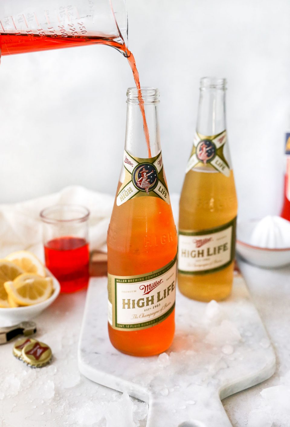 Pouring Aperol liqueur into a Miller High Life bottle to make a Spaghett cocktail. The bottles are on a white surface, next to lemon wedges, a bottle opener & a bottle of Aperol liqueur.