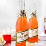 2 Spaghett Cocktails served in Miller High Life bottles. The beer bottles are on a white surface, surrounded by lemon wedges, ice & some Aperol liquor.