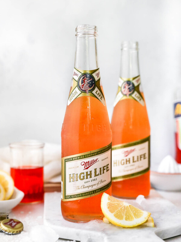 2 Spaghett Cocktails served in Miller High Life bottles. The beer bottles are on a white surface, surrounded by lemon wedges, ice & some Aperol liquor.