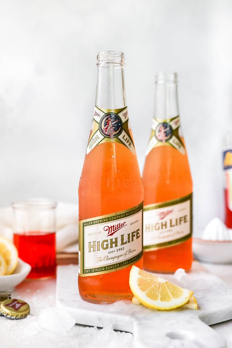 2 Spaghett Cocktails served in Miller High Life bottles. The beer bottles are on a white surface, surrounded by lemon wedges, ice & some Aperol liquor.