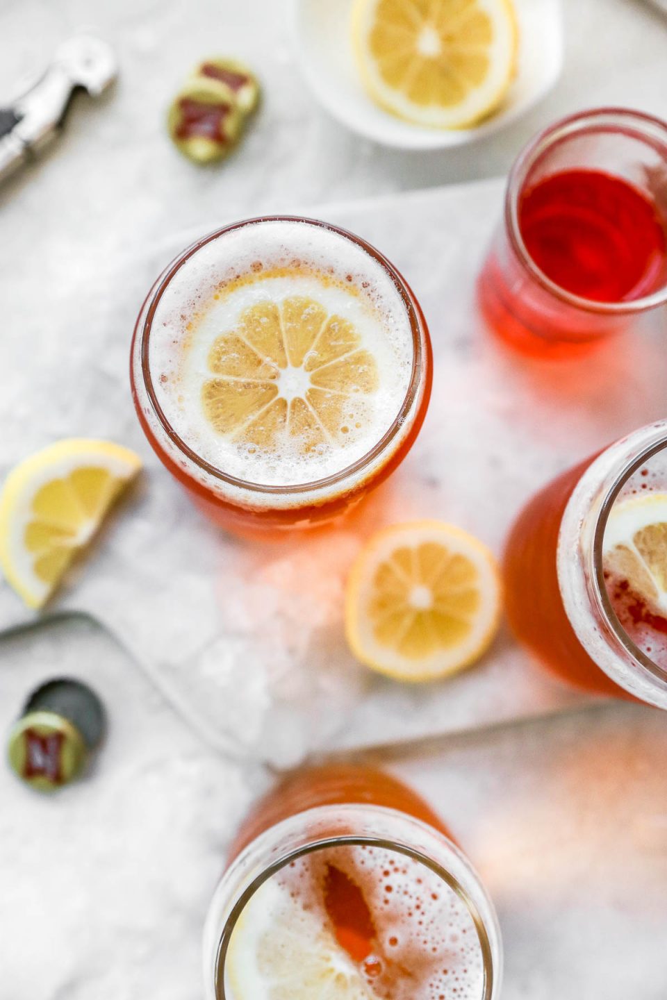Overhead view of 3 spaghett cocktails. The cocktails are in pint glasses, garnished with sliced lemon. Surrounding the glasses are more sliced lemon, Miller High Life bottle caps, a bottle opener, & a small glass jar of bright orange Aperol liqueur.