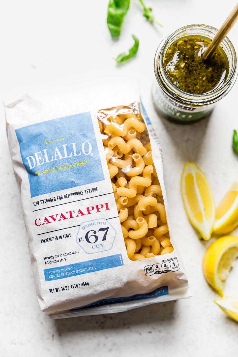 Summer pesto pasta ingredients: DeLallo Cavatappi, DeLallo simply pesto, on a white surface, surrounded by some fresh basil leaves & lemon wedges.