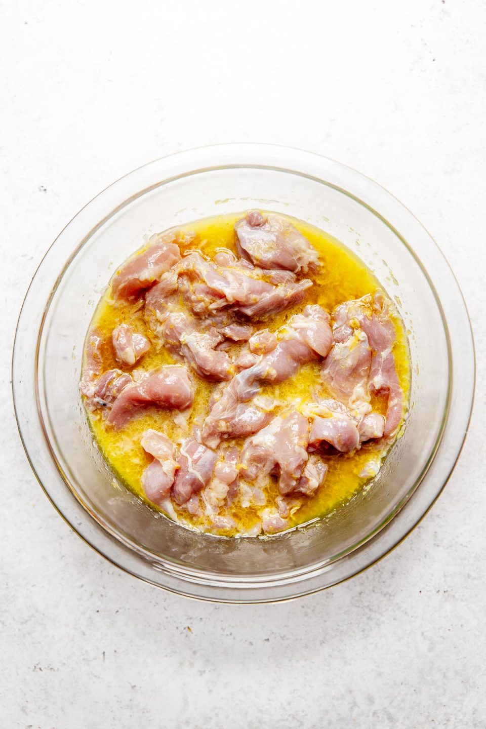 Chicken thighs marinating in a lemon-garlic marinade in a large bowl atop a white surface.