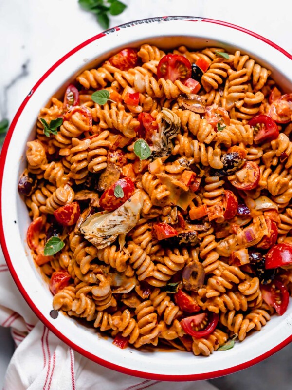 Creamy Italian pasta salad served in a large, white enamel bowl with a cherry red rim. The pasta salad is topped with fresh cherry tomatoes & some oregano leaves. The bowl sits atop a white marble surface, next to a red & white striped linen & some fresh oregano leaves.