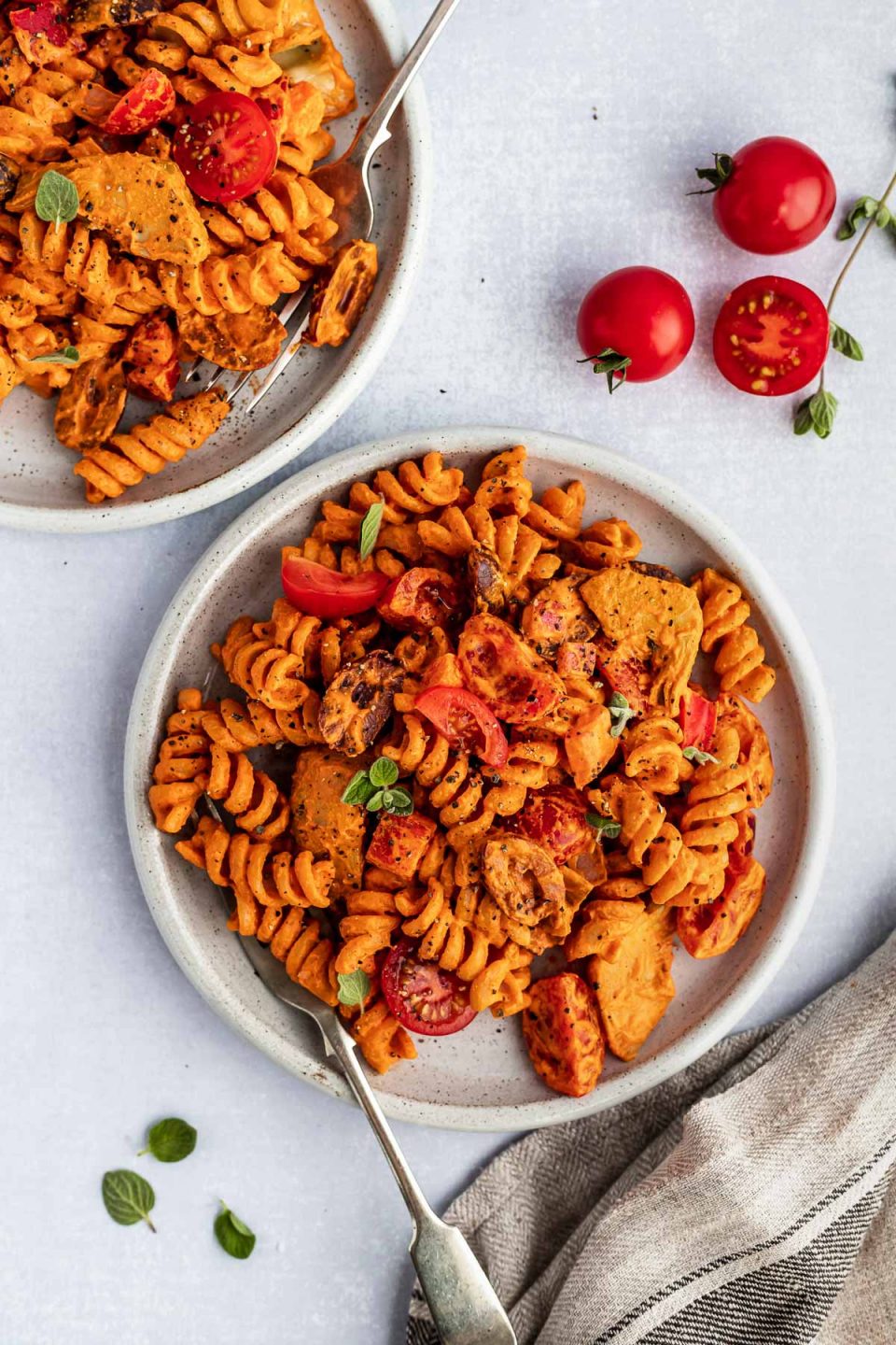 2 individual plates of creamy Italian pasta salad, sitting atop a light blue surface next to a striped linen napkin. The plates are surrounded by fresh oregano & red cherry tomatoes.