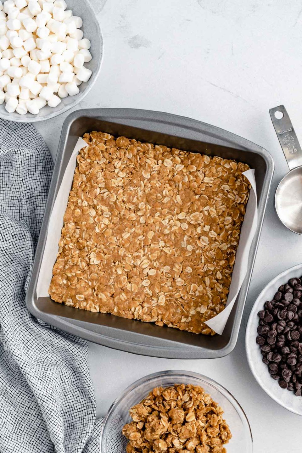 Graham cracker crumb crust pressed into a parchment-lined baking pan. The pan sits atop a white surface, surrounded by small bowls of marshmallows, dark chocolate chips & graham cracker crumb topping.