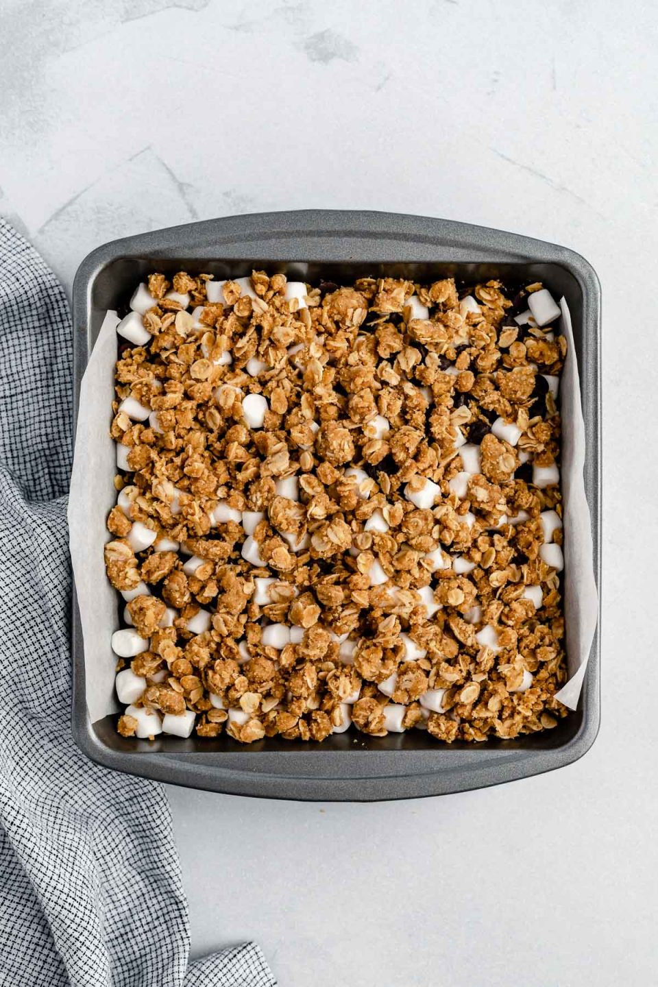 Gooey S'mores Crumble Bars prepped in a parchment-lined baking pan before being baked. The pan sits atop a white surface, with a checkered linen napkin to its side.