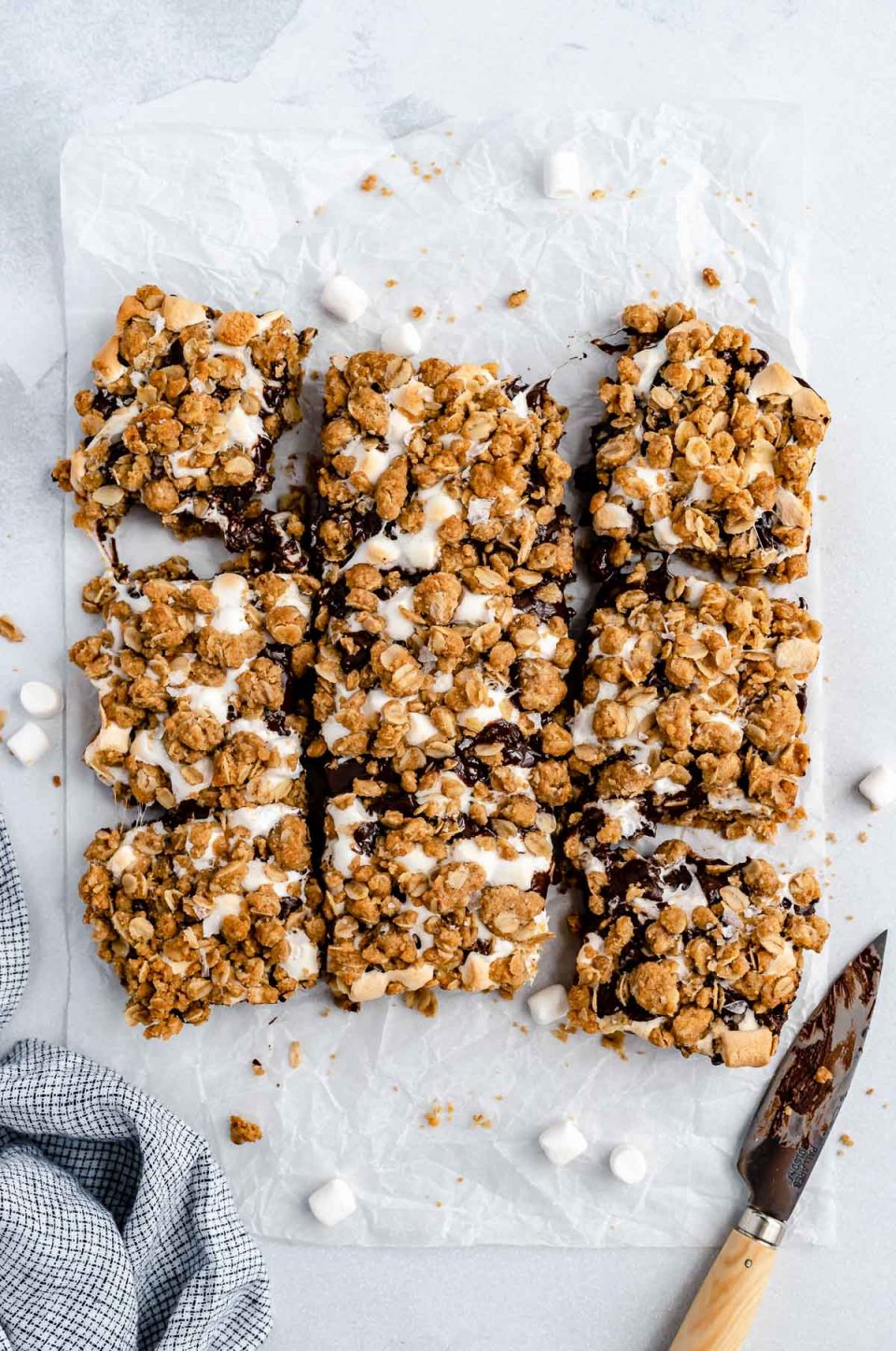 Sliced gooey s'mores crumble bars arranged on a piece of parchment paper atop a white surface. Crumbs & marshmallows are scattered around the bars, as well as a small paring knife that's covered in melted dark chocolate from slicing the bars.