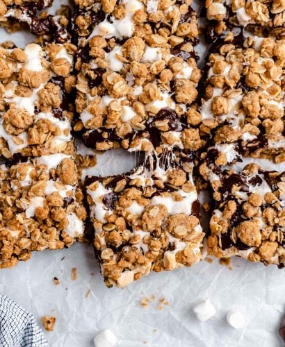 Sliced gooey s'mores crumble bars arranged on a piece of parchment paper atop a white surface. The bars are pulled apart, ever so slightly, with gooey bits of marshmallow strung between the sliced bars.