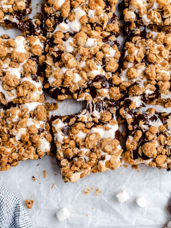 Sliced gooey s'mores crumble bars arranged on a piece of parchment paper atop a white surface. The bars are pulled apart, ever so slightly, with gooey bits of marshmallow strung between the sliced bars.