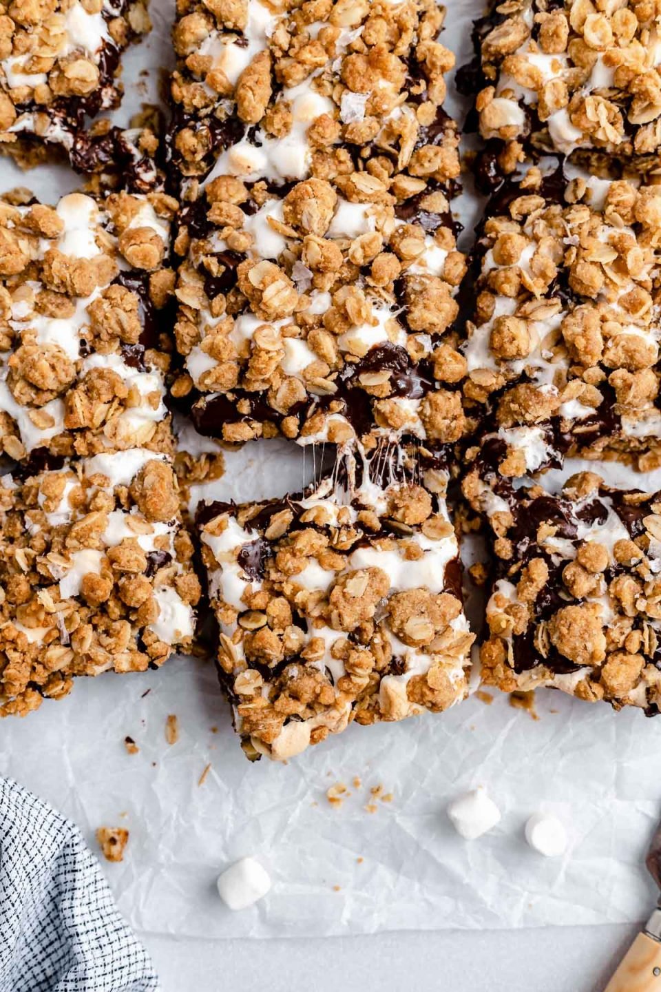 Sliced gooey s'mores crumble bars arranged on a piece of parchment paper atop a white surface. The bars are pulled apart, ever so slightly, with gooey bits of marshmallow strung between the sliced bars.