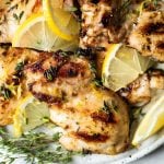 Grilled lemon chicken thighs arranged on a serving plate, with sliced lemon & sprigs of fresh thyme. The plate sits atop a white surface, next to a light blue & white striped linen napkin & a few more sprigs of fresh thyme.