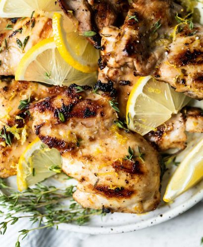 Grilled lemon chicken thighs arranged on a serving plate, with sliced lemon & sprigs of fresh thyme. The plate sits atop a white surface, next to a light blue & white striped linen napkin & a few more sprigs of fresh thyme.