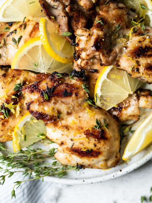 Grilled lemon chicken thighs arranged on a serving plate, with sliced lemon & sprigs of fresh thyme. The plate sits atop a white surface, next to a light blue & white striped linen napkin & a few more sprigs of fresh thyme.
