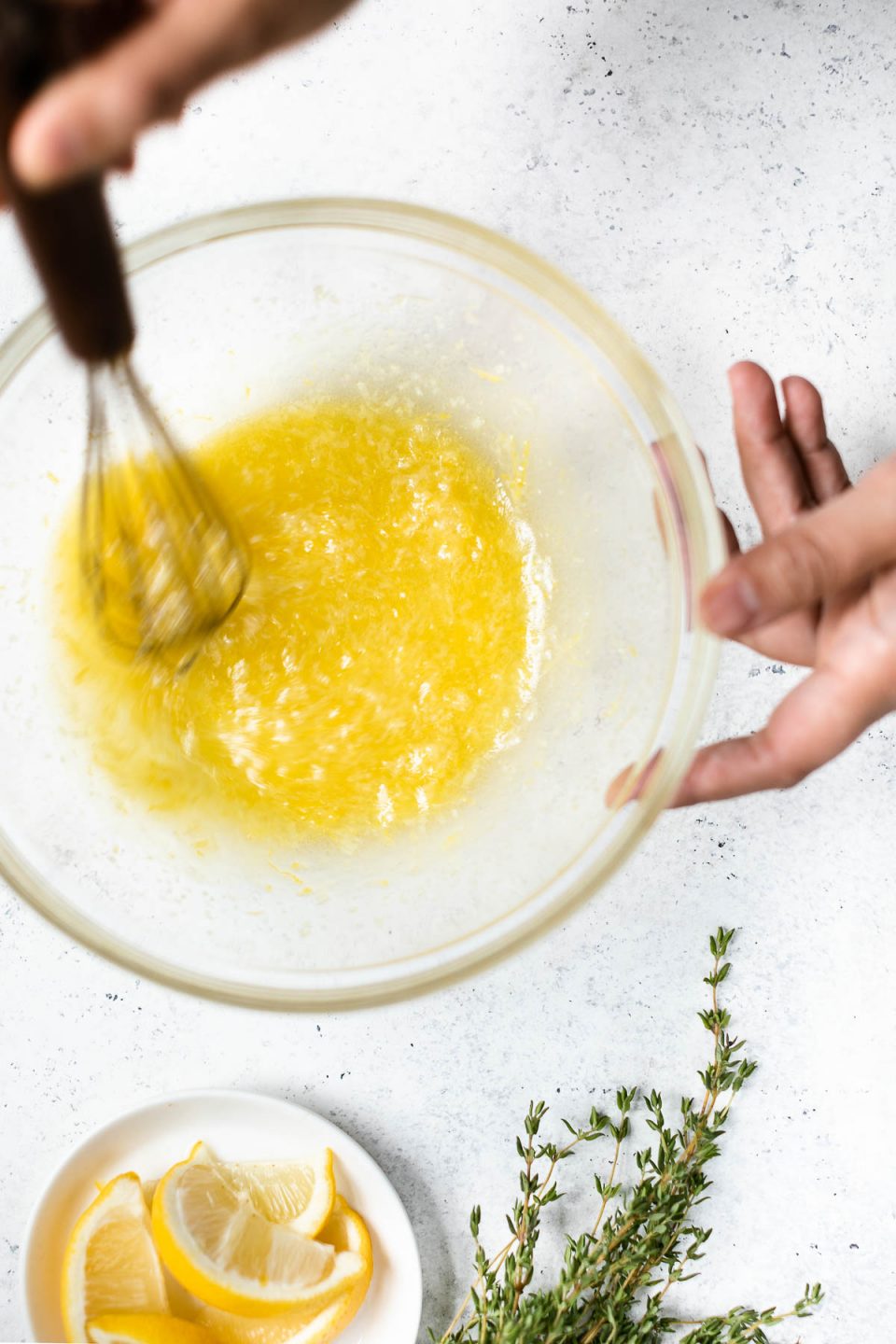A woman's hands shown whisking the lemon garlic marinade together in a glass mixing bowl. The bowl sits atop a white surface, with some lemon wedges & sprigs of fresh thyme next to it. 