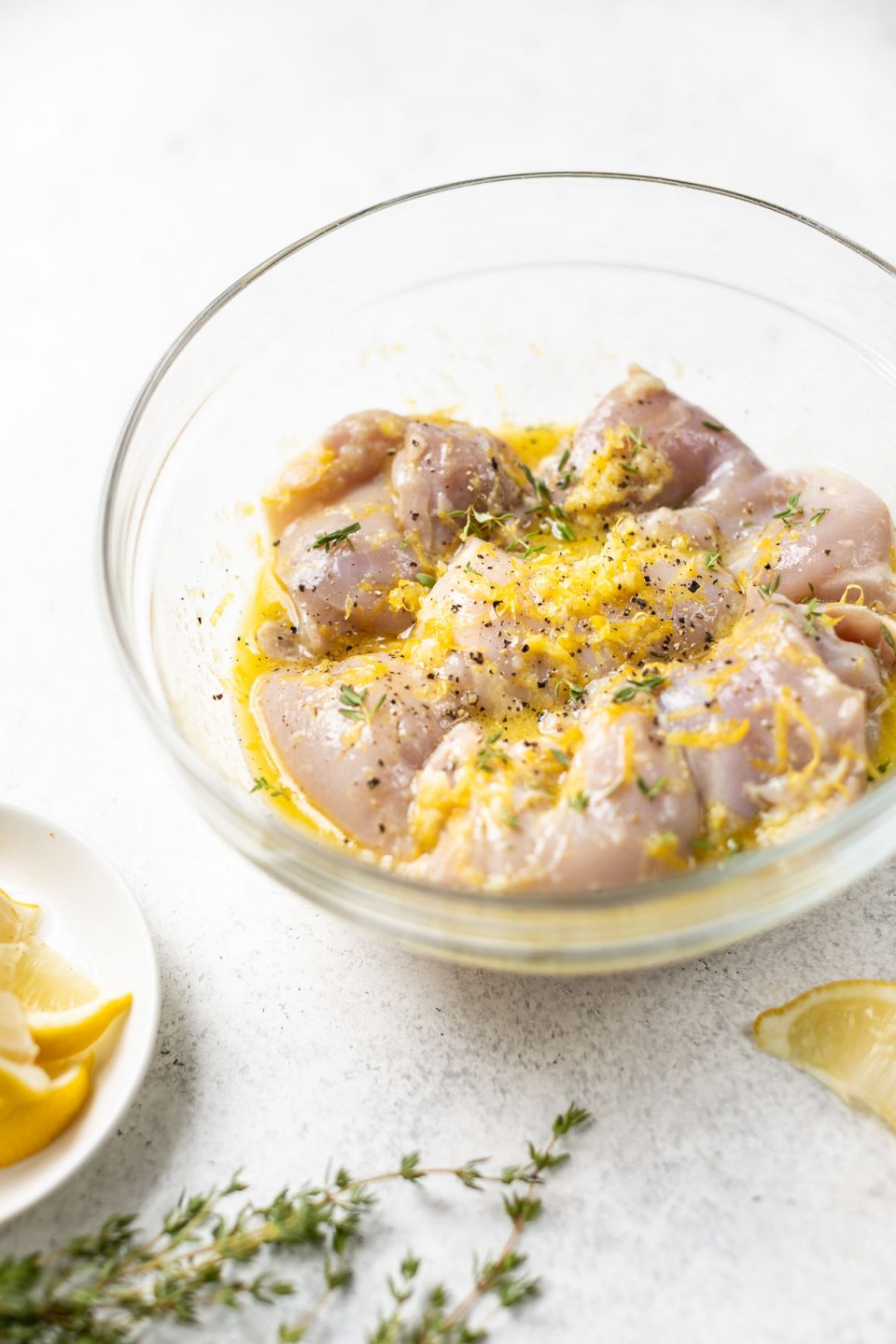 Side angle of chicken thighs marinating in the lemon garlic marinade in a glass mixing bowl. The mixing bowl sits atop a white surface with lemon wedges & sprigs of fresh thyme.
