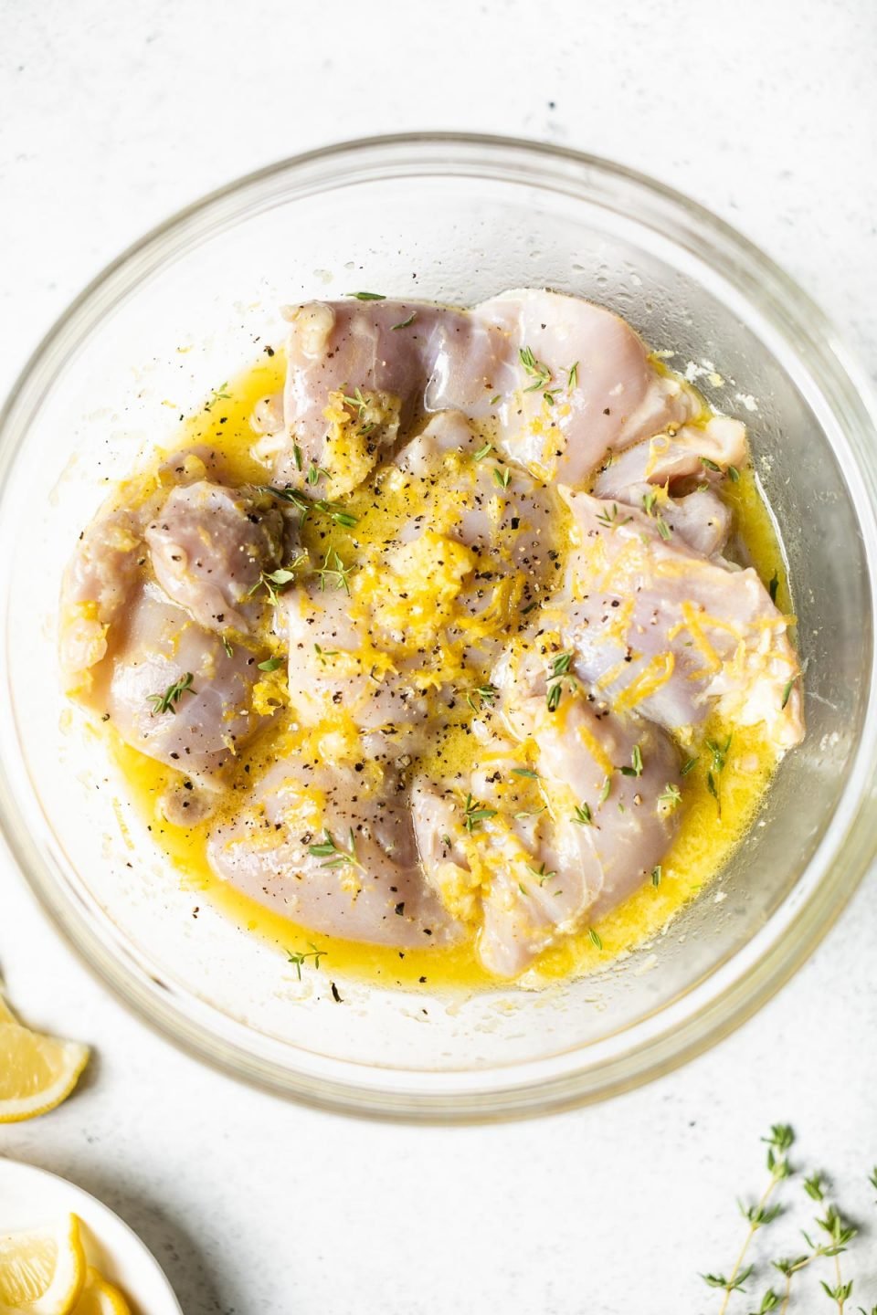 Chicken thighs marinating in the lemon garlic marinade in a glass mixing bowl. The mixing bowl sits atop a white surface with lemon wedges & sprigs of fresh theyme.