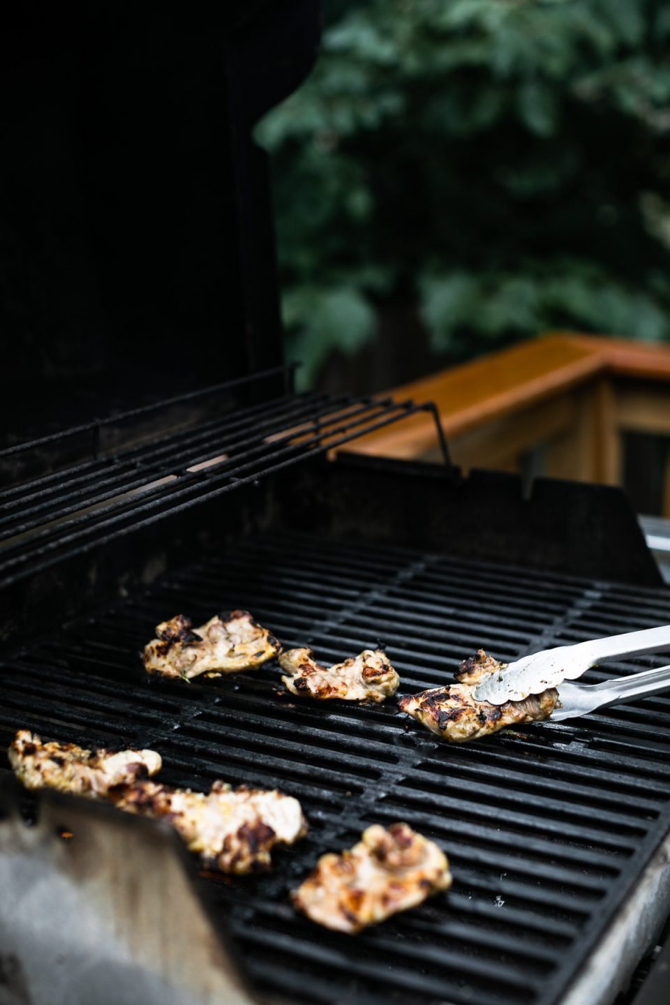 Lemon chicken thighs cooking on the grill. The chicken thighs are directly on grill grates of a gas grill. Tongs poke into the side of the frame, flipping one of the chicken thighs over.