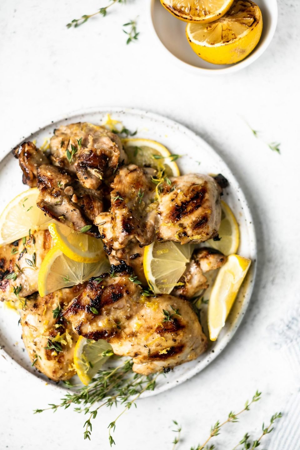Grilled lemon chicken thighs arranged on a serving plate, with sliced lemon & sprigs of fresh thyme. The plate sits atop a white surface, next to a bowl of grilled lemon halves & some sprigs of fresh thyme.