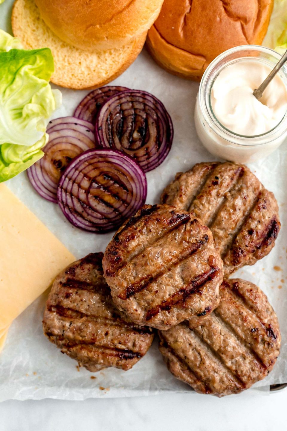 Grilled turkey burgers arranged on a platter with toppings - sliced cheese, leaf lettuce, grilled red onions & a jar of burger sauce.