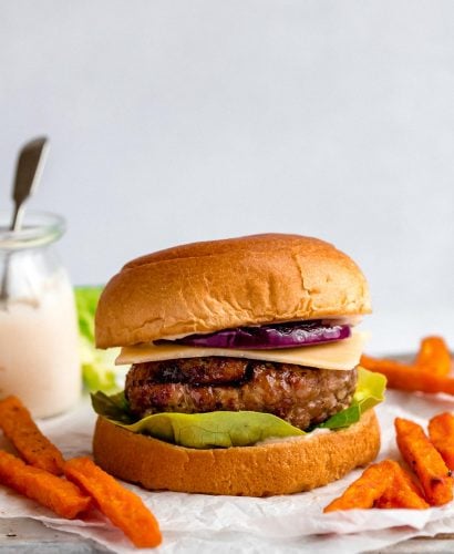 A grilled turkey burger served in a burger bun with lettuce, cheese & grilled red onion. Surrounding the burgers are sweet potato fries, & a small jar of burger sauce.