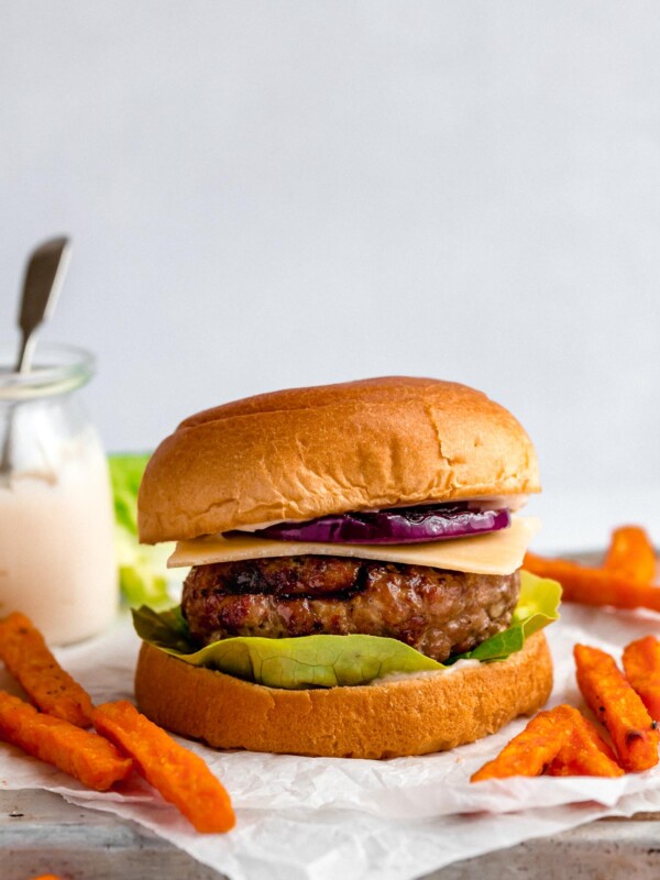 A grilled turkey burger served in a burger bun with lettuce, cheese & grilled red onion. Surrounding the burgers are sweet potato fries, & a small jar of burger sauce.