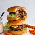 A stack of two grilled turkey burgers, the top one with a bite taken out of it. Surrounding the burgers are sweet potato fries, & a small jar of burger sauce.