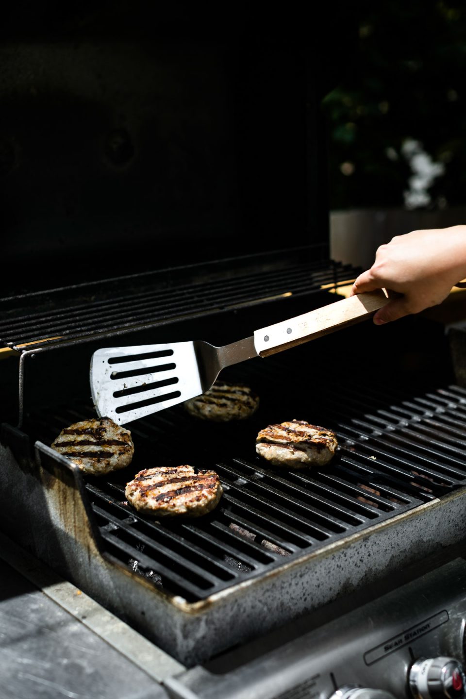 How to grill turkey burgers - 4 turkey burgers being cooked on a gas grill. A woman's hand reaches into the photo with a grill spatula to flip one of the burgers.