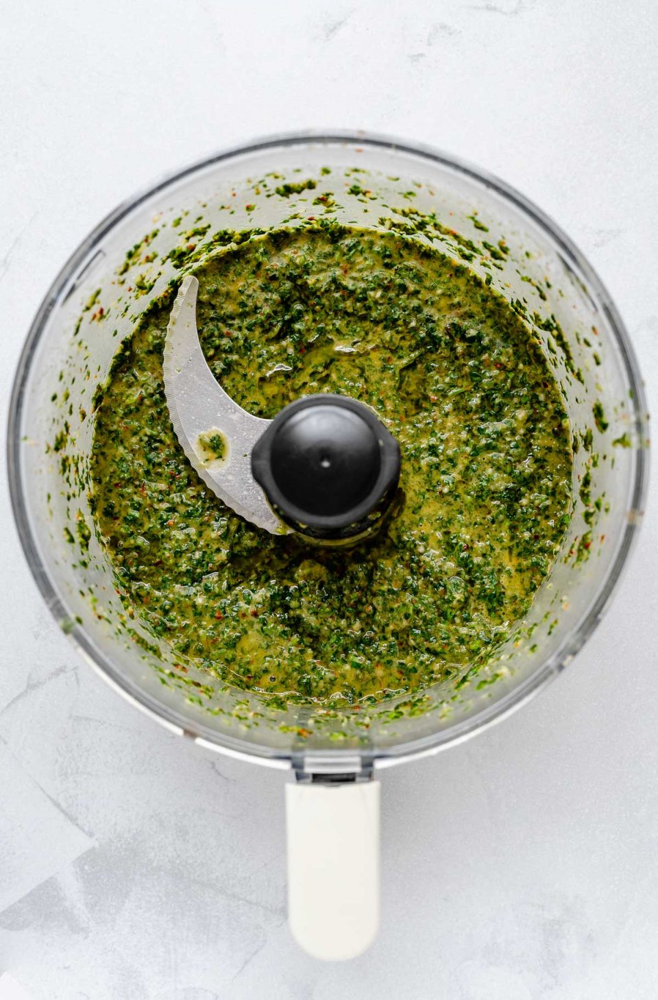 A top down photo of easy Chimichurri sauce that has been pulsed in a food processor bowl. The bowl sits on a light gray surface.