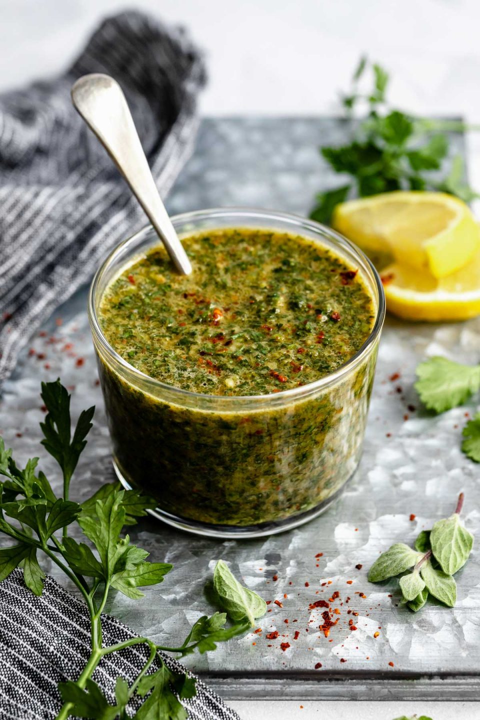 An angled shot of a jar of Chimichurri sauce that sits on a dark gray surface. A spoon has been dipped into the sauce & rests inside the clear glass jar. The jar is surrounded by fresh herbs & a slice of lemon.
