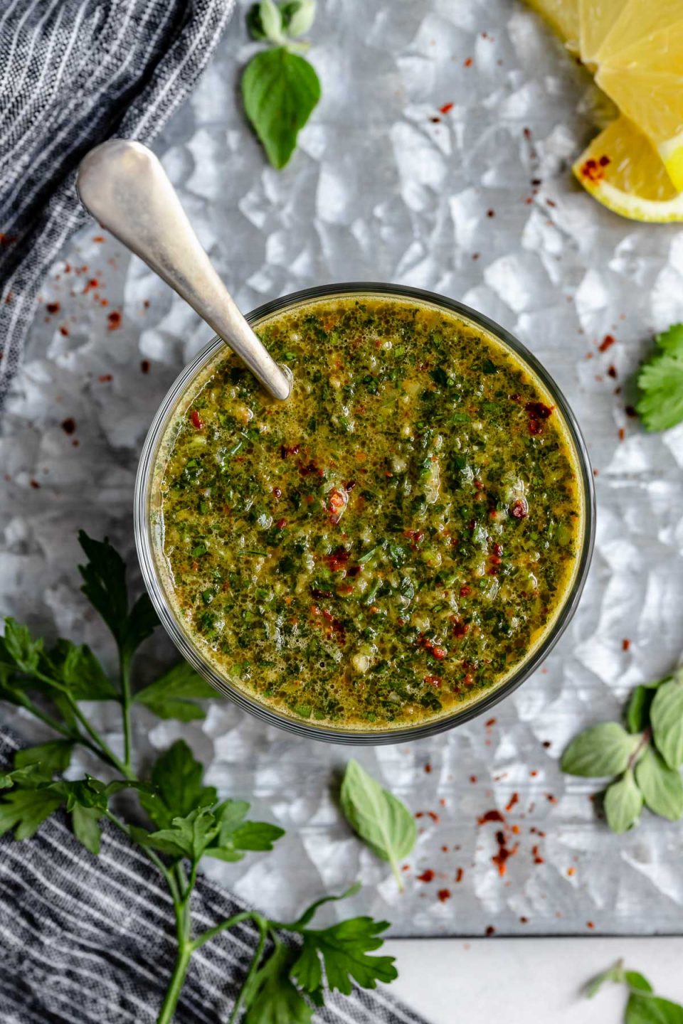 A top down shot of a jar of easy Chimichurri sauce rests on a dark grey surface with fresh herbs and lemons surrounding it. There is a spoon resting inside of the clear glass jar.