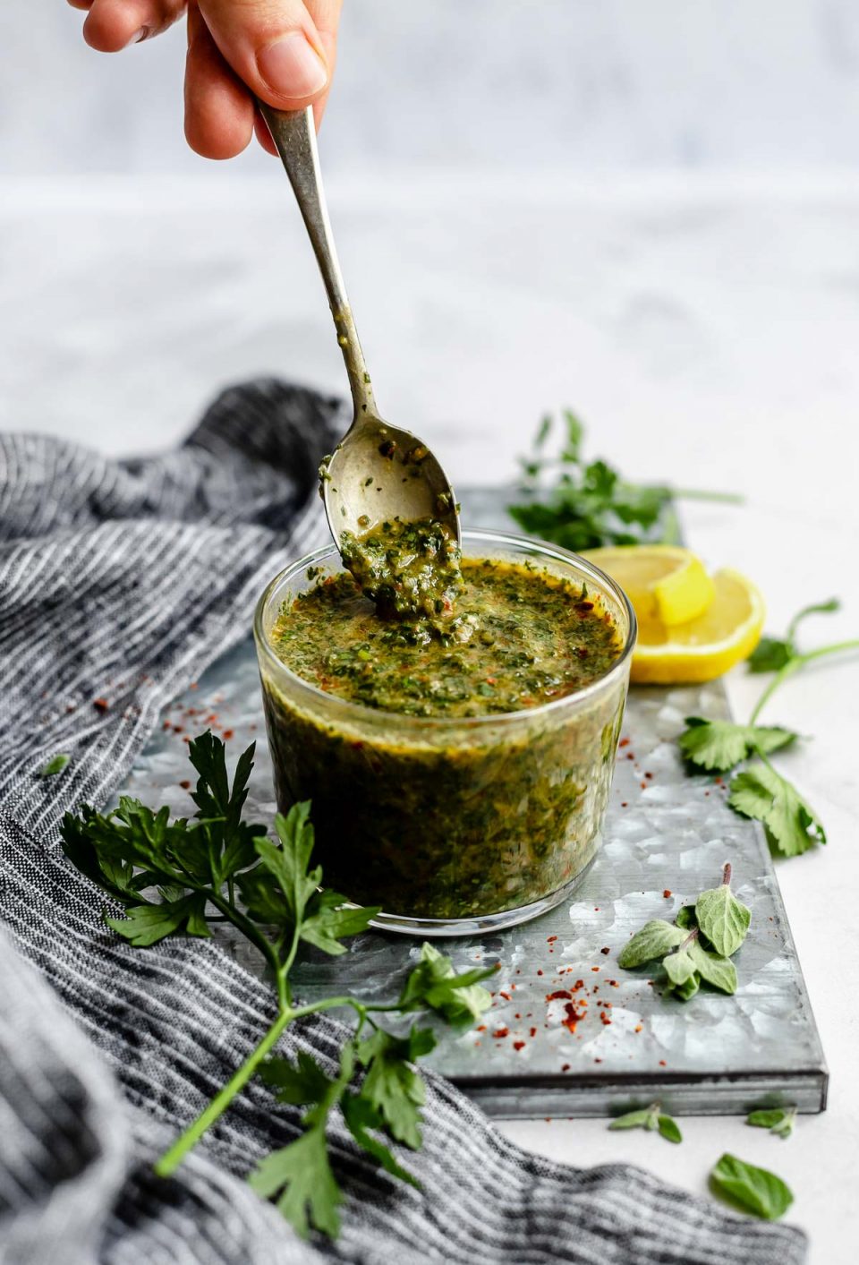 An angled shot of a jar of Chimichurri sauce that sits on a dark gray surface. A spoon has been dipped into the sauce & is being lifted out of the jar with sauce dripping from it. The jar is surrounded by fresh herbs & a slice of lemon.