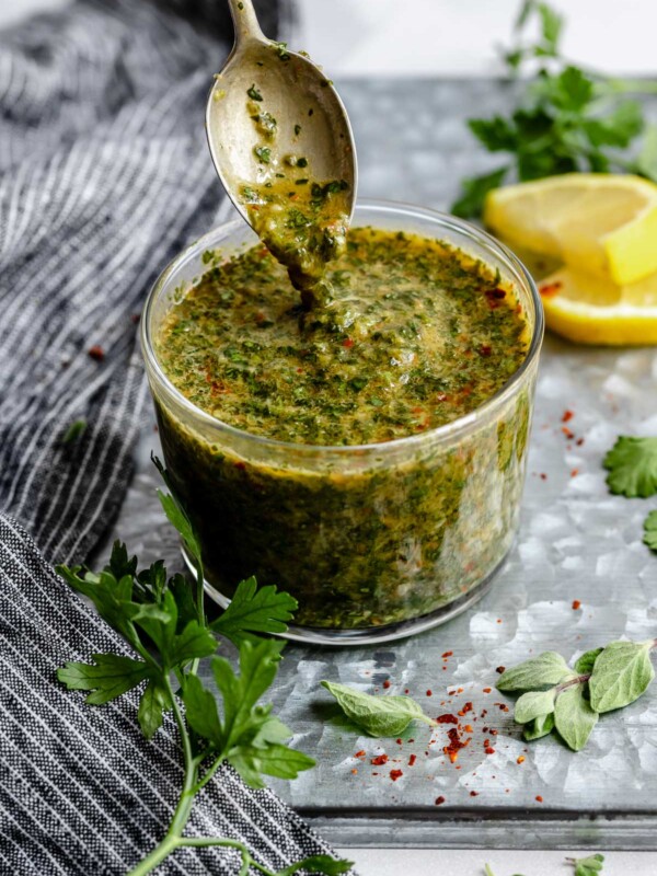 An angled shot of a jar of Chimichurri sauce that sits on a dark gray surface. A spoon has been dipped into the sauce & is being lifted out of the jar with sauce dripping from it. The jar is surrounded by fresh herbs & a slice of lemon.