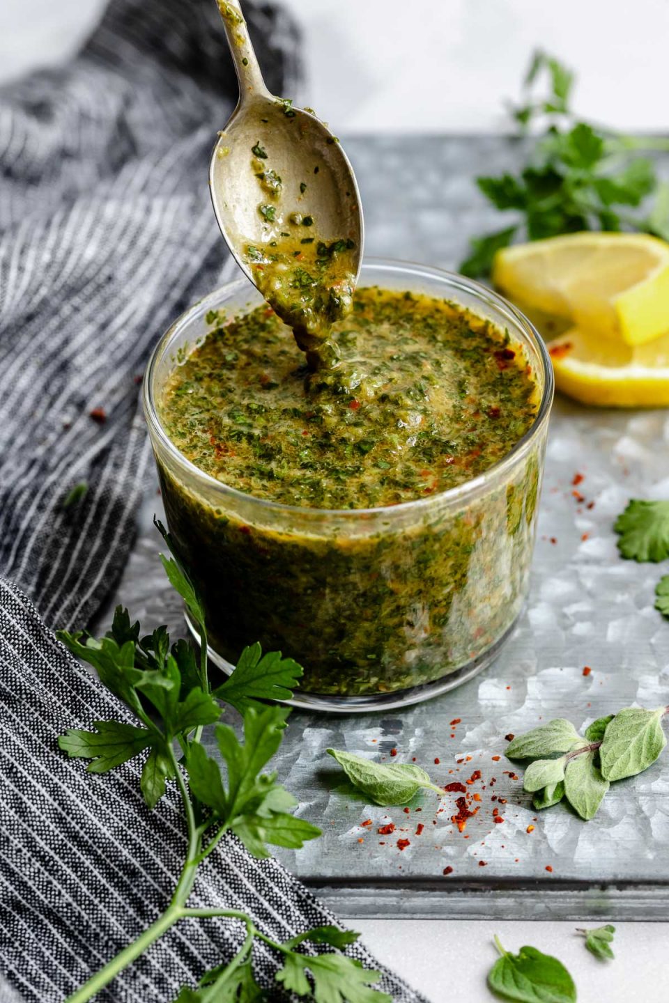 An angled shot of a jar of Chimichurri sauce that sits on a dark gray surface. A spoon has been dipped into the sauce & is being lifted out of the jar with sauce dripping from it. The jar is surrounded by fresh herbs & a slice of lemon.