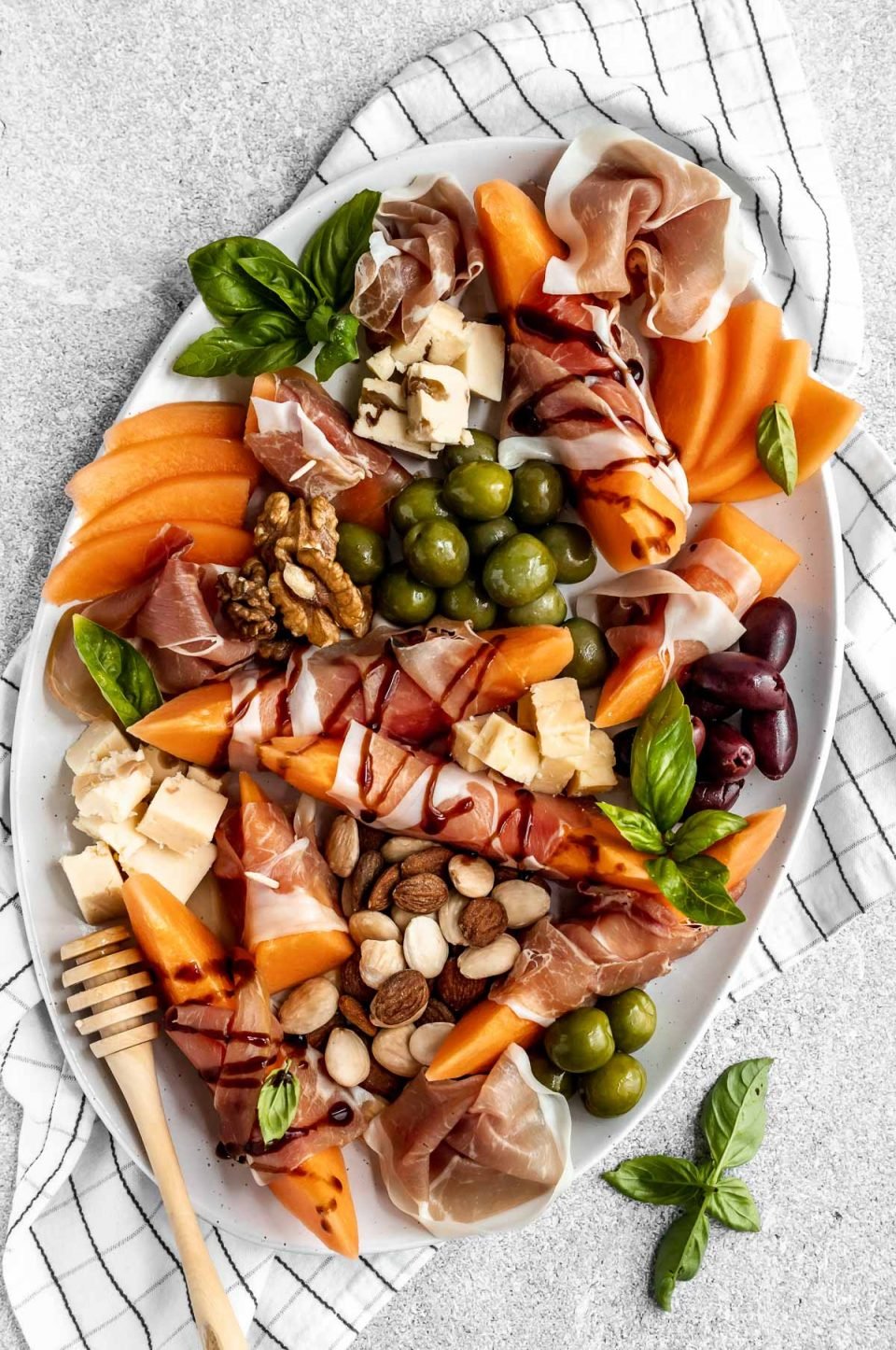 Prosciutto and melon cheese board! Prosciutto-Wrapped Cantaloupe on a large serving platter with olives, nuts, cheese, & fresh basil leaves. Some pieces of prosciutto-wrapped melon are drizzled with balsamic syrup.