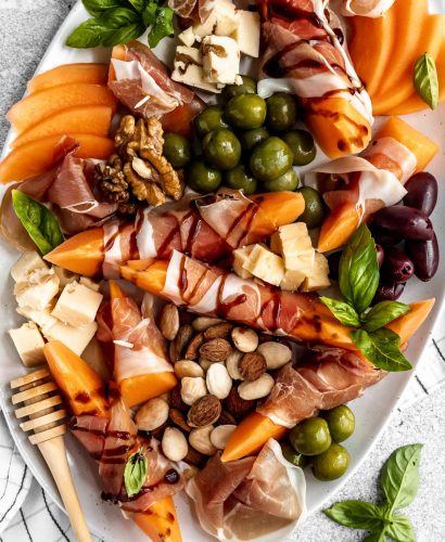 Close up of prosciutto and melon cheese board! Prosciutto-Wrapped Cantaloupe on a large serving platter with olives, nuts, cheese, & fresh basil leaves. Some pieces of prosciutto-wrapped melon are drizzled with balsamic syrup.