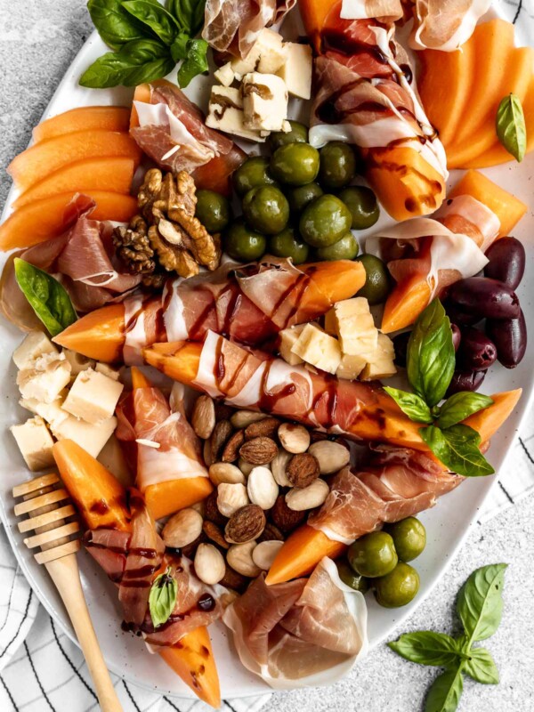 Close up of prosciutto and melon cheese board! Prosciutto-Wrapped Cantaloupe on a large serving platter with olives, nuts, cheese, & fresh basil leaves. Some pieces of prosciutto-wrapped melon are drizzled with balsamic syrup.