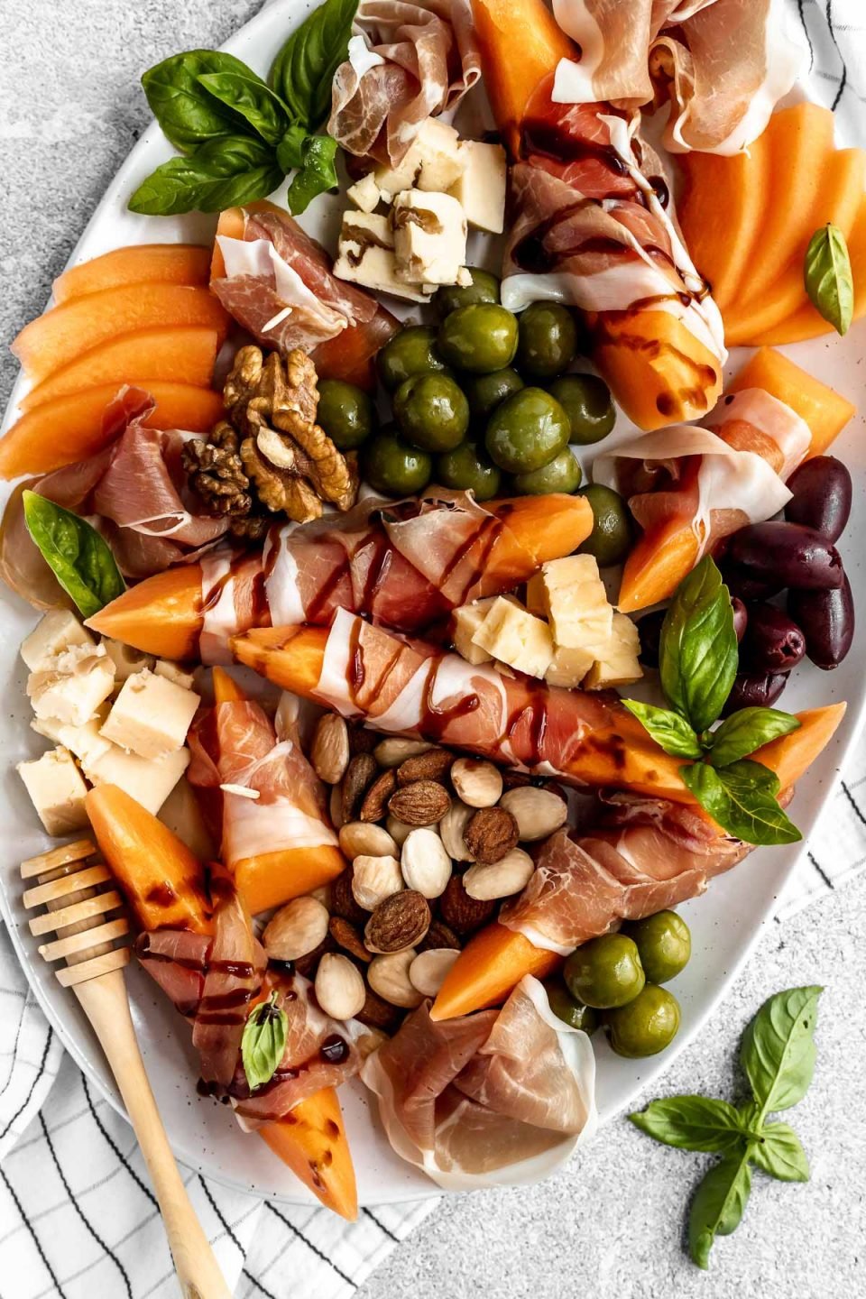 Close up of prosciutto and melon cheese board! Prosciutto-Wrapped Cantaloupe on a large serving platter with olives, nuts, cheese, & fresh basil leaves. Some pieces of prosciutto-wrapped melon are drizzled with balsamic syrup.