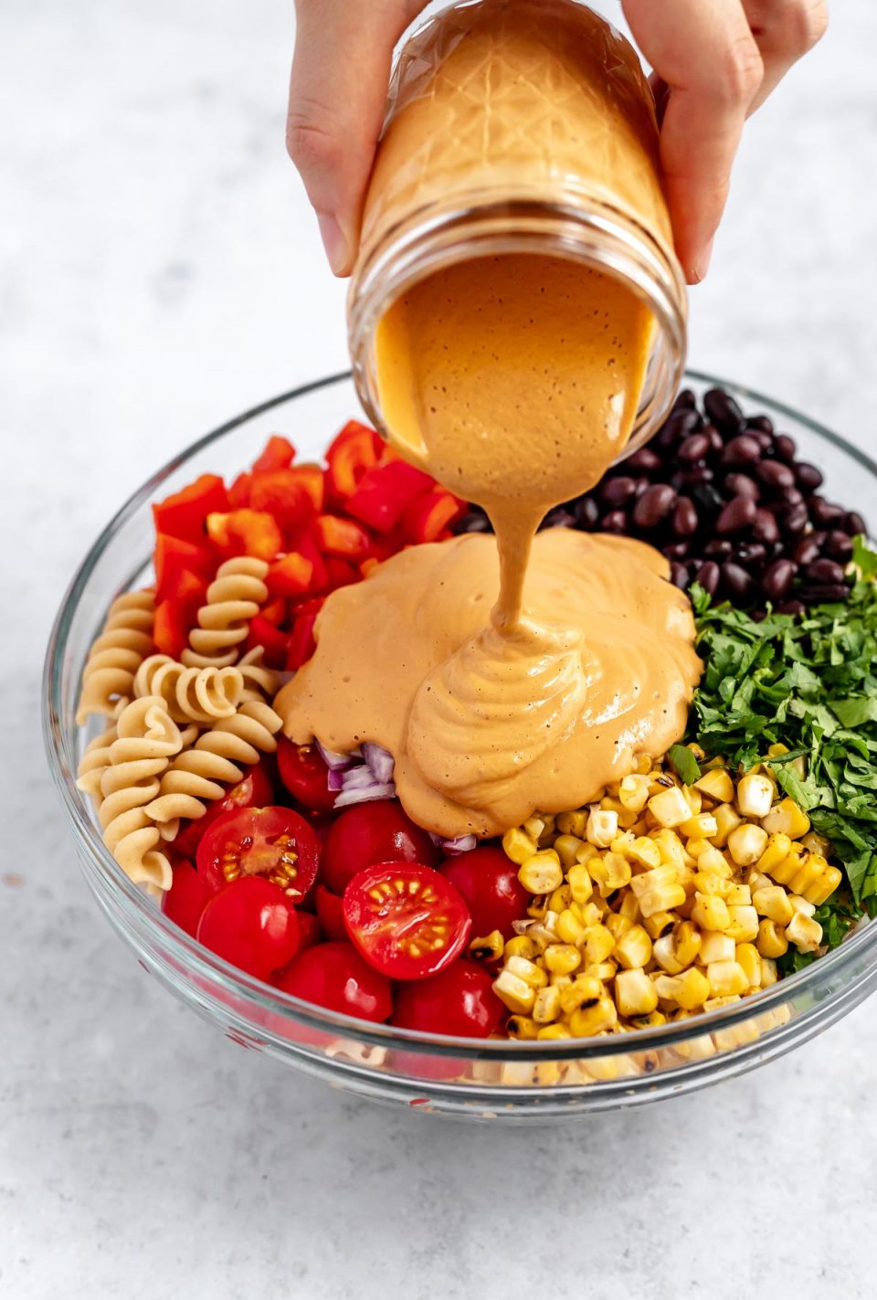 A woman's hand pouring the creamy vegan Southwest dressing over the Southwest Pasta Salad in a large mixing bowl. 