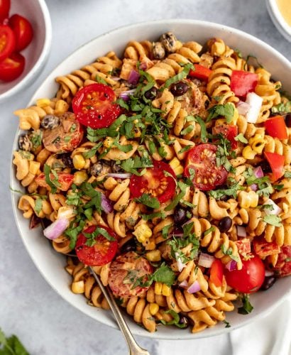 Creamy Vegan Southwest Pasta Salad shown in a large white serving bowl. The salad is garnished with freshly chopped cilantro. The bowl is placed on a light blue surface, surrounded with smaller white dishes filled with tomatoes, chopped cilantro, & creamy vegan southwest dressing.