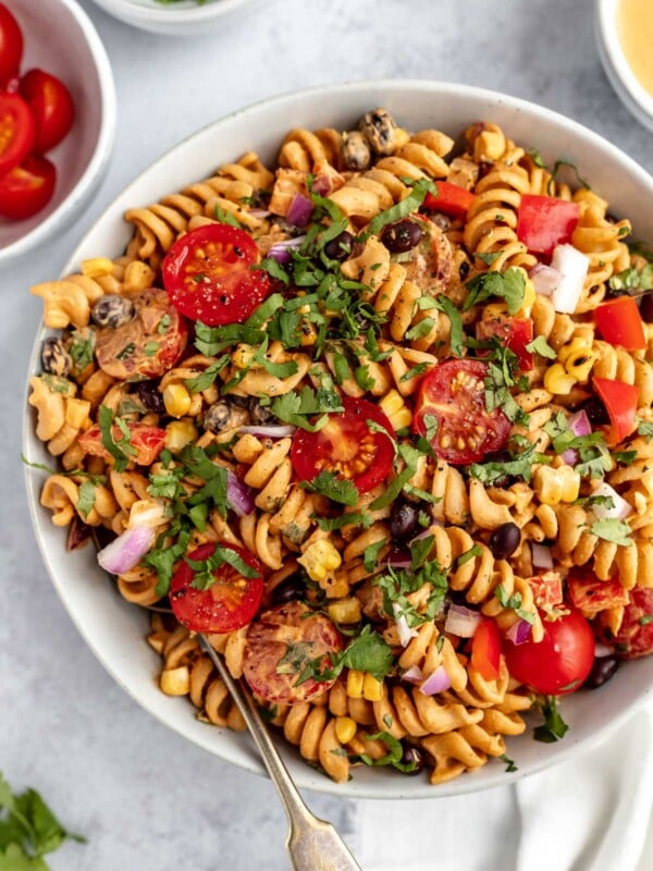 Creamy Vegan Southwest Pasta Salad shown in a large white serving bowl. The salad is garnished with freshly chopped cilantro. The bowl is placed on a light blue surface, surrounded with smaller white dishes filled with tomatoes, chopped cilantro, & creamy vegan southwest dressing.