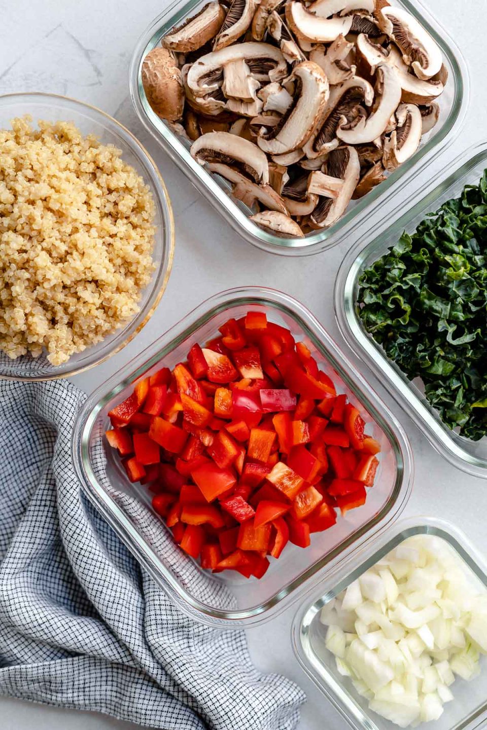 Chicken sausage quinoa skillet ingredients arranged on a creamy white textured surface: varied sizes of glass meal prep containers are filled with cooked quinoa, thinly sliced baby portobello mushrooms, finely shredded. kale, diced red bell pepper, and diced yellow onion. A blue and white plaid linen napkin rests alongside the open meal prep containers.