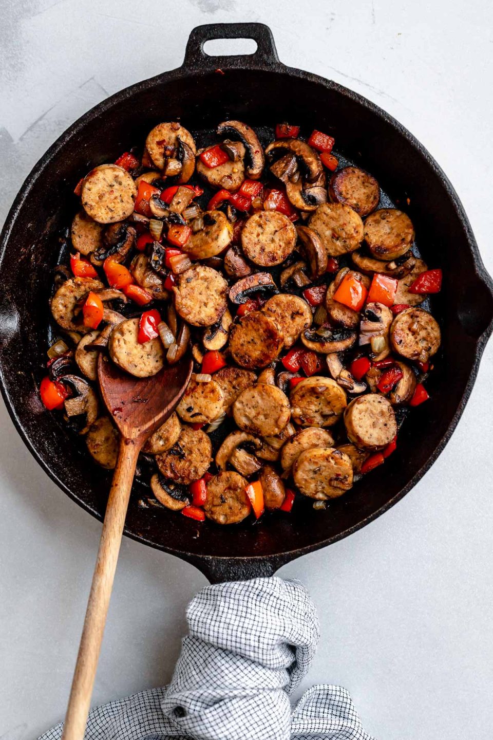 How to make Italian chicken sausage and quinoa skillet, Step 1 & 2: cook the aromatics & brown the sausage and mushrooms. A large black cast iron skillet sits atop a creamy white textured surface and is filled with softened onion and bell peppers, browned Italian chicken sausage and baby portobello mushrooms. A wooden spoon used for stirring rests inside of the skillet and a blue and white plaid linen napkin is wrapped around the skillet handle.