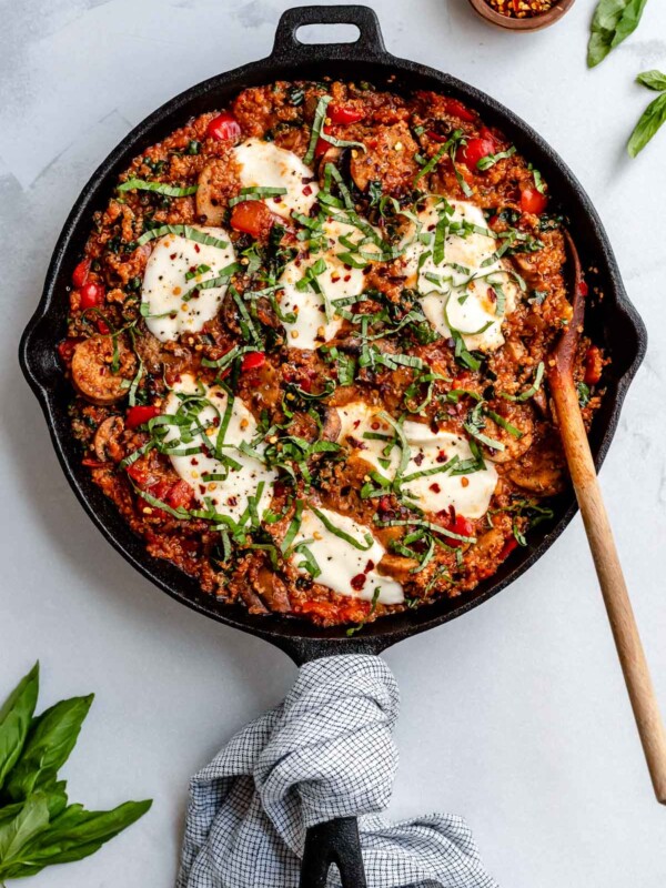 An overhead shot of a finished Italian chicken sausage and quinoa skillet fills a large black cast iron skillet that sits atop a creamy white textured surface. Melted fresh mozzarella tops the skillet dinner with crushed red chili flakes, thinly sliced basil, and grated parmesan used for garnish. A small wooden pinch bowl filled with crushed red chili flakes and loose fresh basil leaves surround the skillet. A blue and white plaid linen napkin is tied around the skillet handle and a wooden serving spoon rests inside of the skillet.
