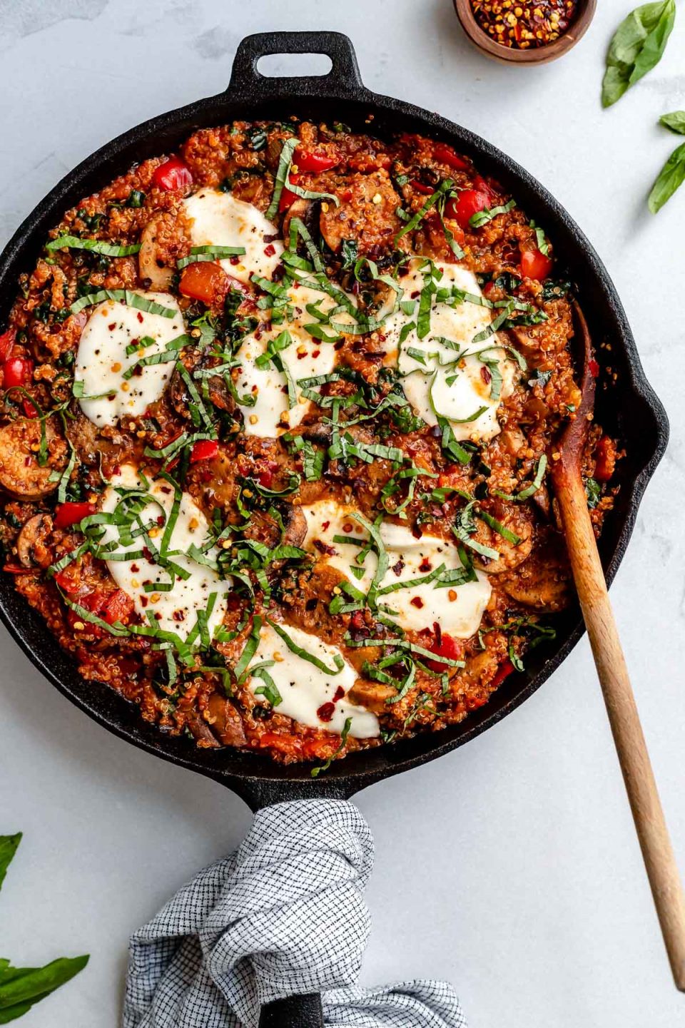 An overhead shot of a finished Italian chicken sausage and quinoa skillet fills a large black cast iron skillet that sits atop a creamy white textured surface. Melted fresh mozzarella tops the skillet dinner with crushed red chili flakes, thinly sliced basil, and grated parmesan used for garnish. A small wooden pinch bowl filled with crushed red chili flakes and loose fresh basil leaves surround the skillet. A blue and white plaid linen napkin is tied around the skillet handle and a wooden serving spoon rests inside of the skillet.