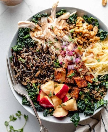 A large white bowl filled with autumn grain bowl ingredients: shredded kale, wild rice, quinoa, apple, roasted sweet potato, diced red onion, shredded cheese, chopped walnuts & shredded chicken. A fork is placed in the grain bowl, nestled to the left side under the wild rice & quinoa. The bowl sits atop a light blue surface, surrounded by sprigs of fresh thyme & a striped linen napkin. Next to the bowl are some whole walnuts & a jar of maple balsamic dressing.