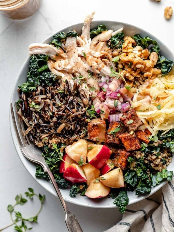A large white bowl filled with autumn grain bowl ingredients: shredded kale, wild rice, quinoa, apple, roasted sweet potato, diced red onion, shredded cheese, chopped walnuts & shredded chicken. A fork is placed in the grain bowl, nestled to the left side under the wild rice & quinoa. The bowl sits atop a light blue surface, surrounded by sprigs of fresh thyme & a striped linen napkin. Next to the bowl are some whole walnuts & a jar of maple balsamic dressing.